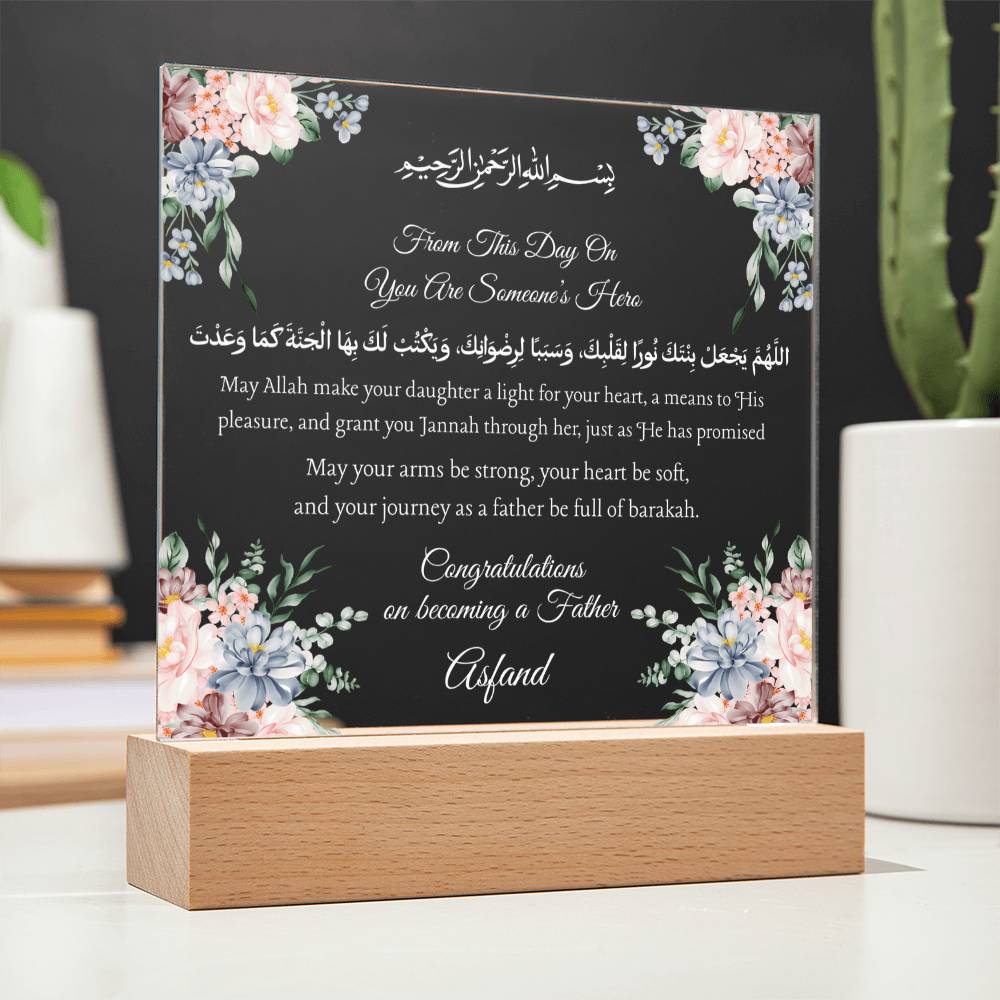 a wooden stand with a clear glass plaque featuring a floral design and Arabic text. The plaque is placed on a white surface, and there is a potted plant in the background.